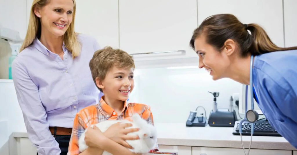 young boy holds a white rabbit at the vet with his mom while talking with the female vet to answer do rabbits need to be spayed or neutered